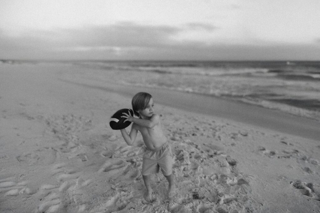 A young boy plays with a football on the white sand beaches of 30A, captured in a timeless black and white family photo that tells a generational story.