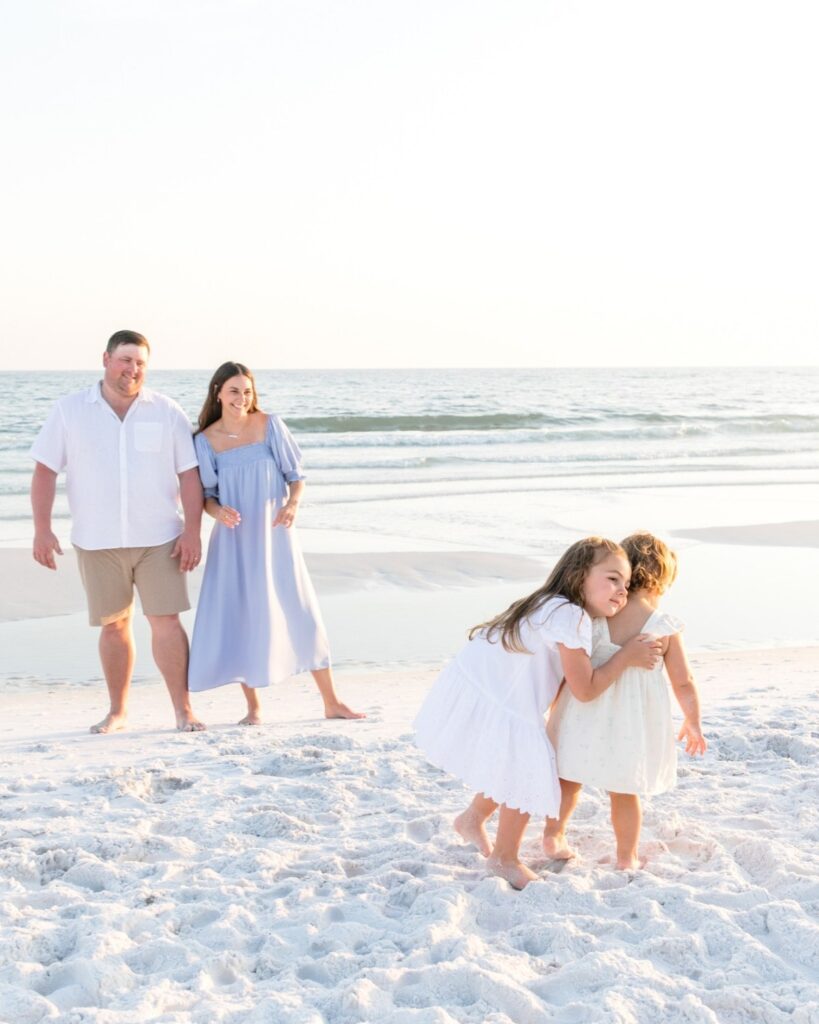 Young sisters playing on the beach while their parents stand nearby during a 30A extended family photography session.