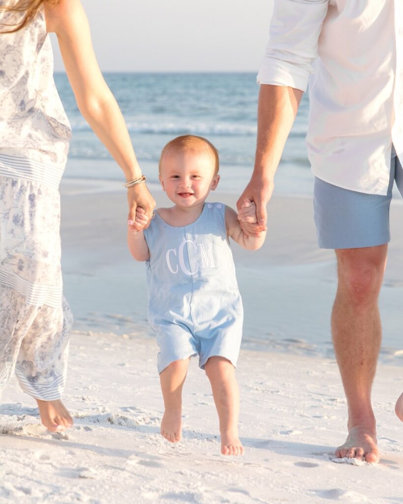 Toddler walking on the beach while holding both parents’ hands during a 30A extended family photography session.