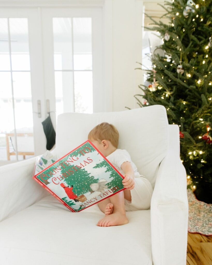 Toddler sibling reading a Christmas book in front of the Christmas tree during an in-home lifestyle newborn session in Santa Rosa Beach Florida