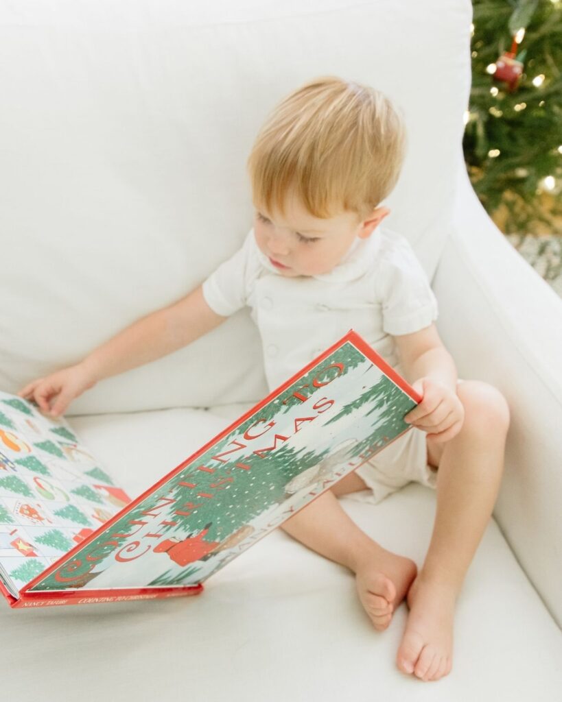 Toddler sibling reading a Christmas book in front of the Christmas tree during an in-home lifestyle newborn session in Santa Rosa Beach Florida
